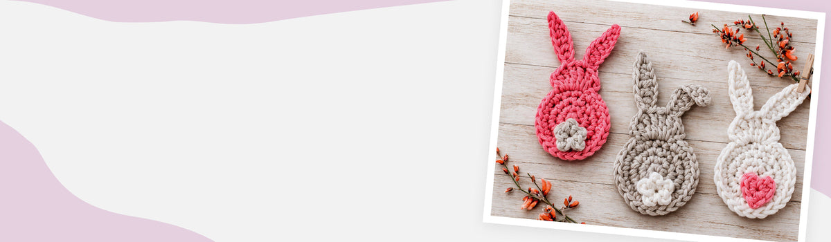 Drei gehäkelte Hasen-Applikationen in Pink, Taupe und Weiß auf beigefarben getünchtem Holz im Vintage-Look, Produktbeispiel für Deko häkeln für Anfänger