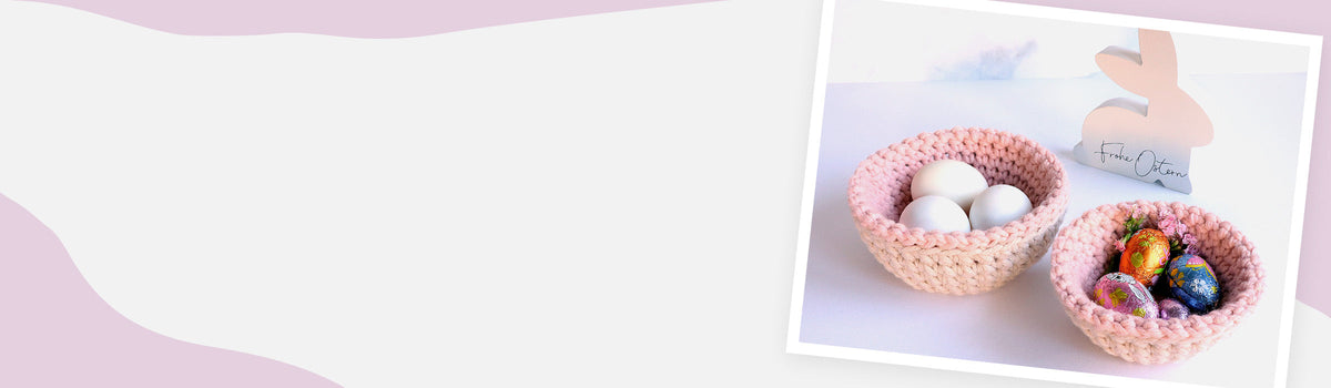 Zwei gehäkelte Nester in Rosa-Beige auf hellem Hintergrund – eines mit weißen Eiern, eines mit bunten Schokoeiern; im Hintergrund ein Osterhase aus Holz mit „Frohe Ostern“