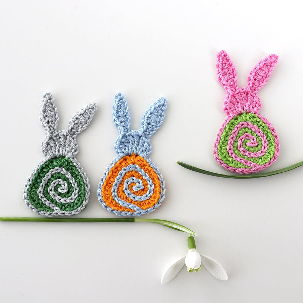 Drei gehäkelte Hasenmotive „Hasi Spirelli“ in Grau, Blau und Pink mit farbigen Spiralen neben einem Schneeglöckchen.