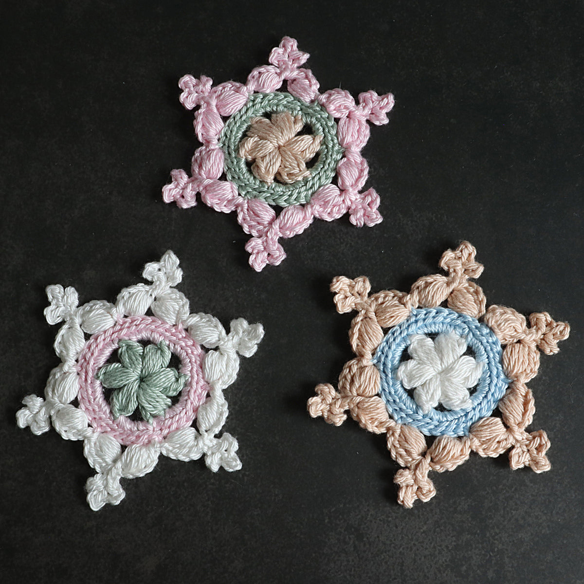 Drei gehäkelte Schneeflocken „Kleine Helena“ in Rosa, Grün, Mint und Beige auf schwarzem Hintergrund.