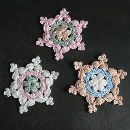 Drei gehäkelte Schneeflocken „Kleine Helena“ in Rosa, Grün, Mint und Beige auf schwarzem Hintergrund.