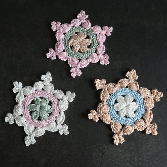 Drei gehäkelte Schneeflocken „Kleine Helena“ in Rosa, Grün, Mint und Beige auf schwarzem Hintergrund.