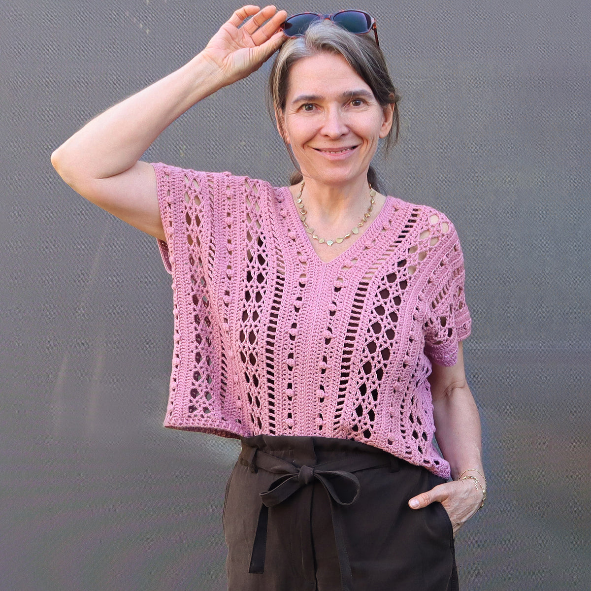 Häkeldesignerin Elke Eder trägt das rosa gehäkelte Shirt Jolanda vor grauem Hintergrund.