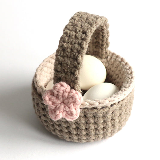 Gehäkelter Henkelkorb in Beige und Braun mit rosa Blume, gefüllt mit weißen Eiern.