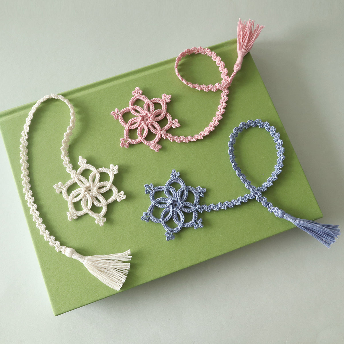 Drei gehäkelte Lesezeichen „Lorelei“ in Rosa, Creme und Blau mit Schneeflocken und Quasten auf einem grünen Buch