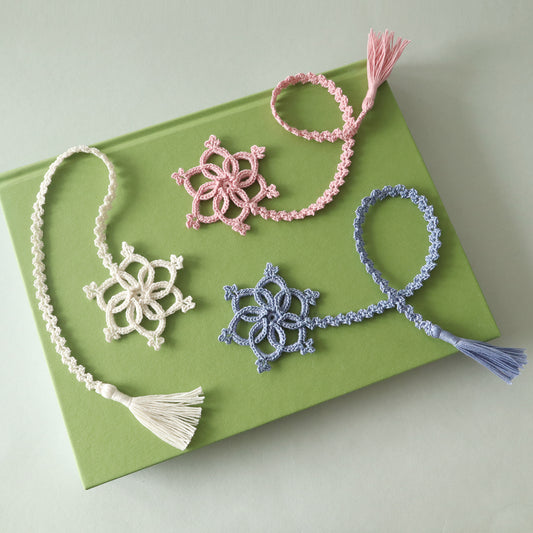Drei gehäkelte Lesezeichen „Lorelei“ in Rosa, Creme und Blau mit Schneeflocken und Quasten auf einem grünen Buch