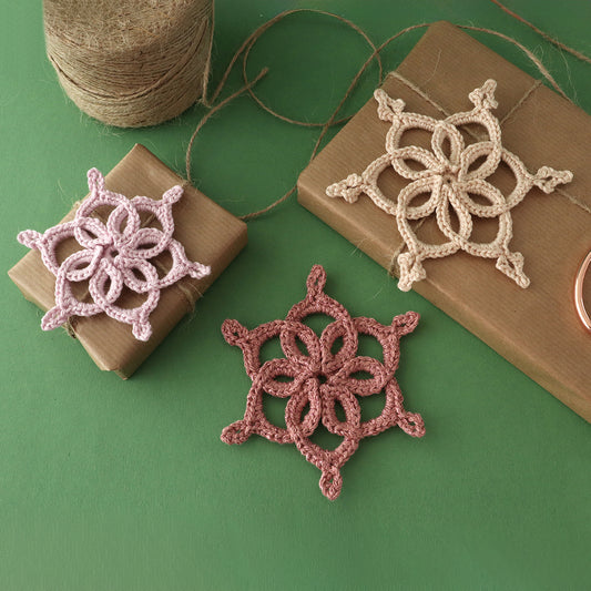 Drei gehäkelte Schneeflocken „Lorelei“ in Rosa, Beige und Altrosa neben verpackten Geschenken auf grünem Untergrund