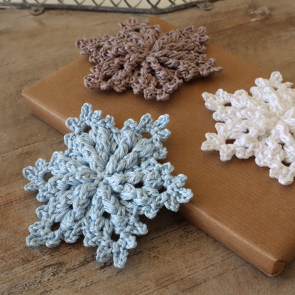 Drei gehäkelte Schneeflocken „Neve“ in Hellblau, Weiß und Taupe mit plastischem Reliefmuster auf einem Geschenkpaket und Holztisch