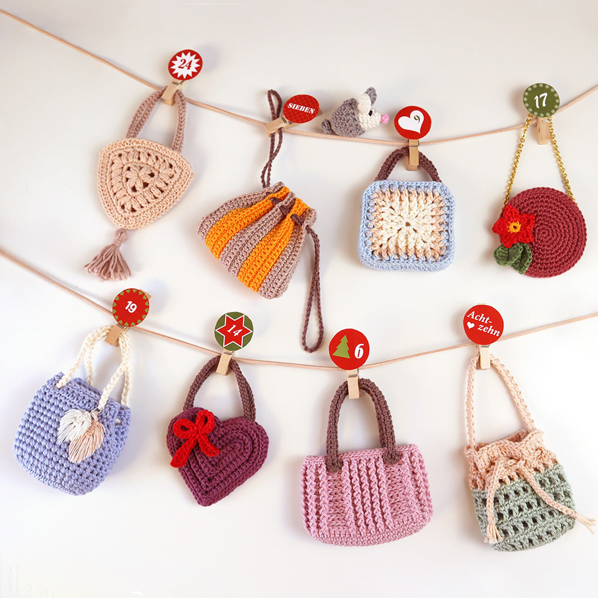 Acht gehäkelte Mini-Taschen des Adventskalenders „Fashionistas 2.0“ in verschiedenen Farben, mit nummerierten Holzklammern an zwei Schnüren aufgehängt