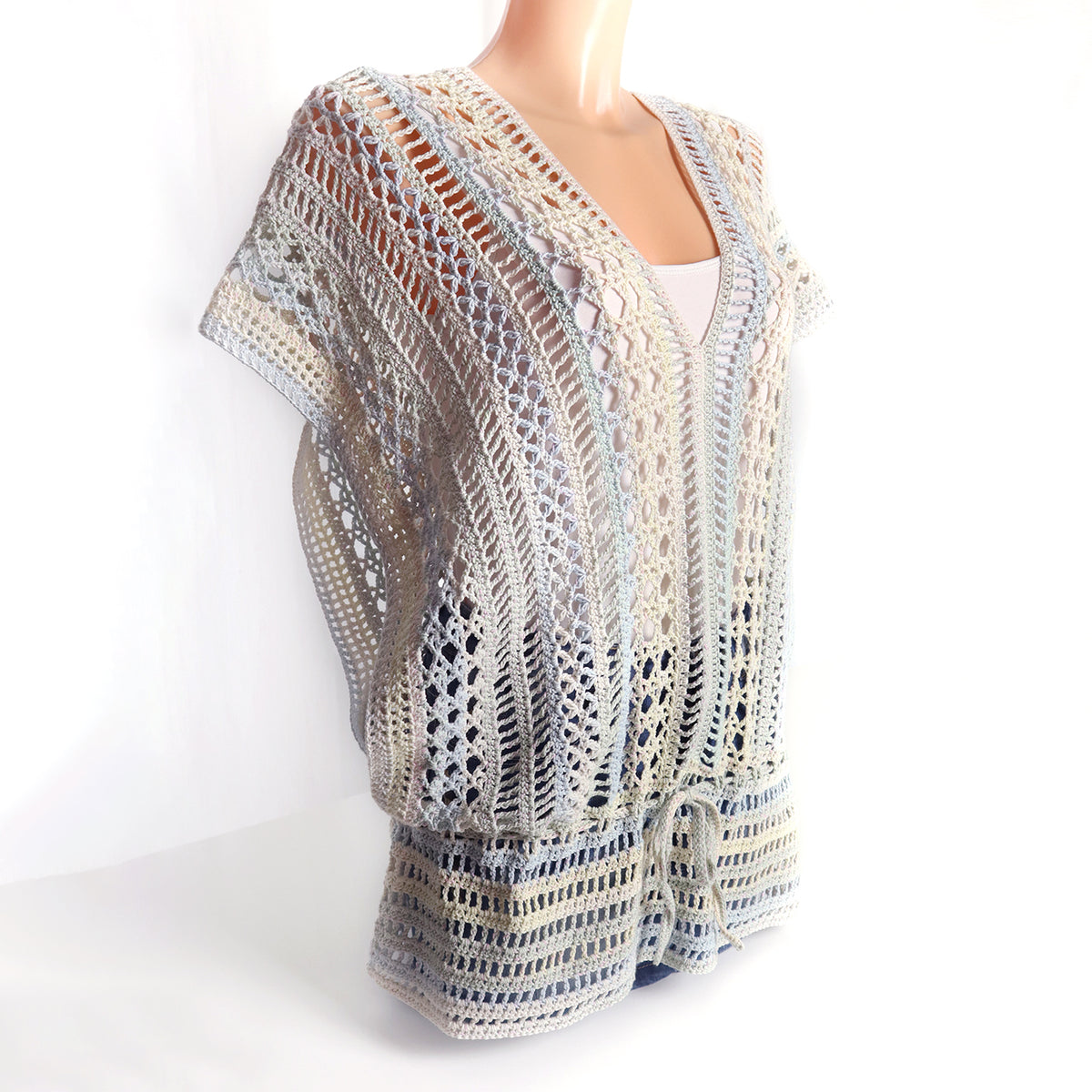 Gehäkelte Tunika „Trinity“ in Creme, Hellblau und Beige mit V-Ausschnitt, luftigem Mustermix und Bindegürtel, seitlich auf einer Schneiderpuppe gezeigt.