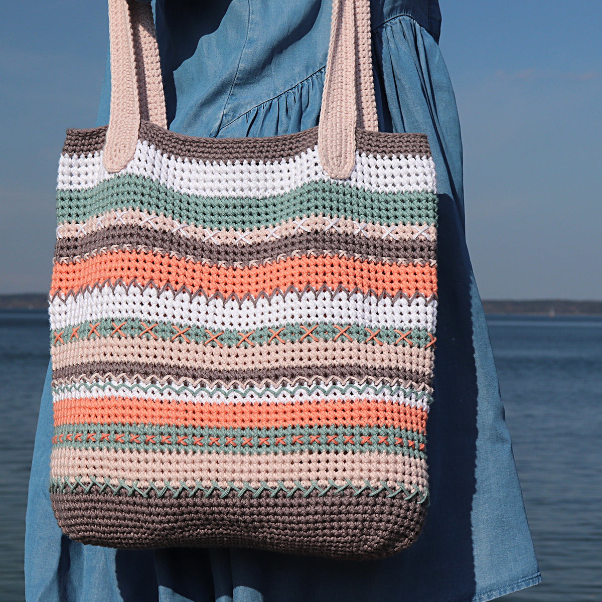 Gehäkelte gestreifte Tasche „Helena“ in Grau, Rosa, Weiß und Mint mit dekorativen Stickereien, getragen vor blauem Hintergrund